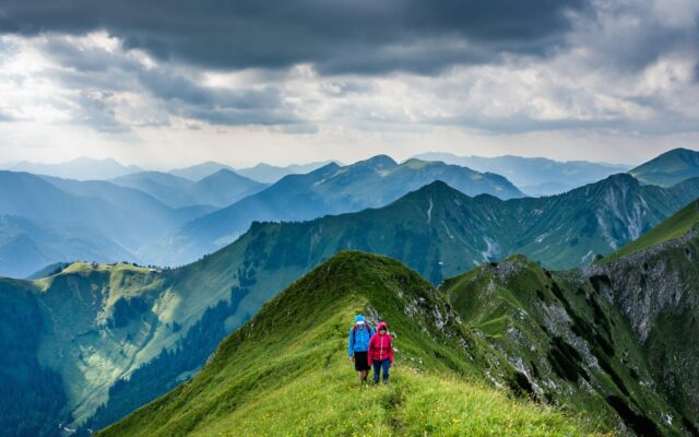 Hai người đi bộ trên sườn núi xanh giữa các dãy núi phía xa